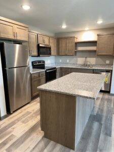 Modern kitchen with wood cabinets, stainless steel appliances including a refrigerator, oven, and microwave. Granite countertops feature prominently on the island and surrounding surfaces, complemented by recessed lighting and wood-patterned flooring.