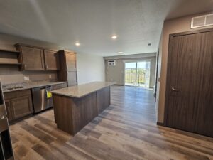 Modern kitchen with wood cabinets, an island, and stainless steel appliances. The open layout extends to a living area with wood flooring and sliding glass doors leading to a balcony with a view of greenery.