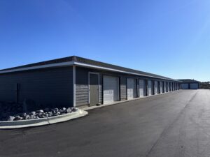 A long row of storage unit garages with closed white doors under a clear blue sky. The building is surrounded by an asphalt driveway and a small rock-covered area to the side.