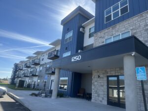 A modern three-story apartment building with a stone and blue exterior. The entrance has the number "1250" above it. Balconies line the upper floors, and a handicapped parking sign is visible in the foreground.