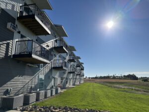 A row of modern apartment balconies faces a sunny, open field. The sky is clear and bright, with the sun casting long shadows on the ground. The landscape is grassy and flat, extending toward a line of trees in the distance.